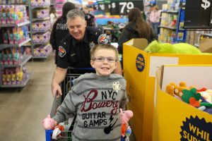 A young boy in a grey sweater smiles with a Bannock County Sheriff's deputy while shopping at WalMart. The child sits in a shopping cart that the deputy pushes behind him