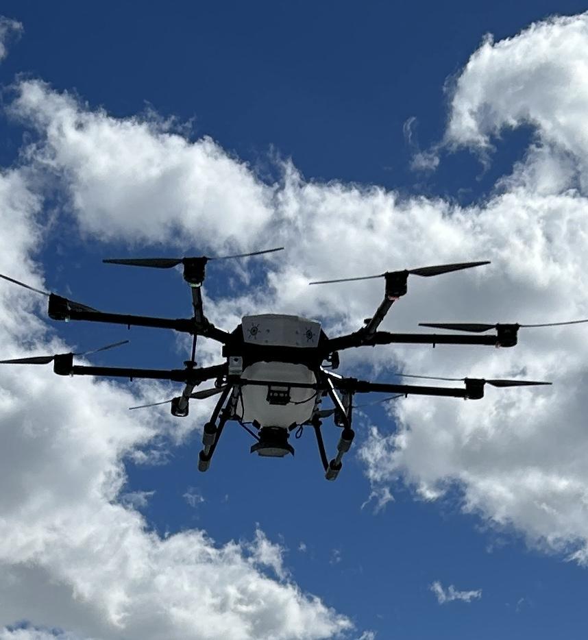a drone with six propellers flying with a blue sky and white clouds in the background.